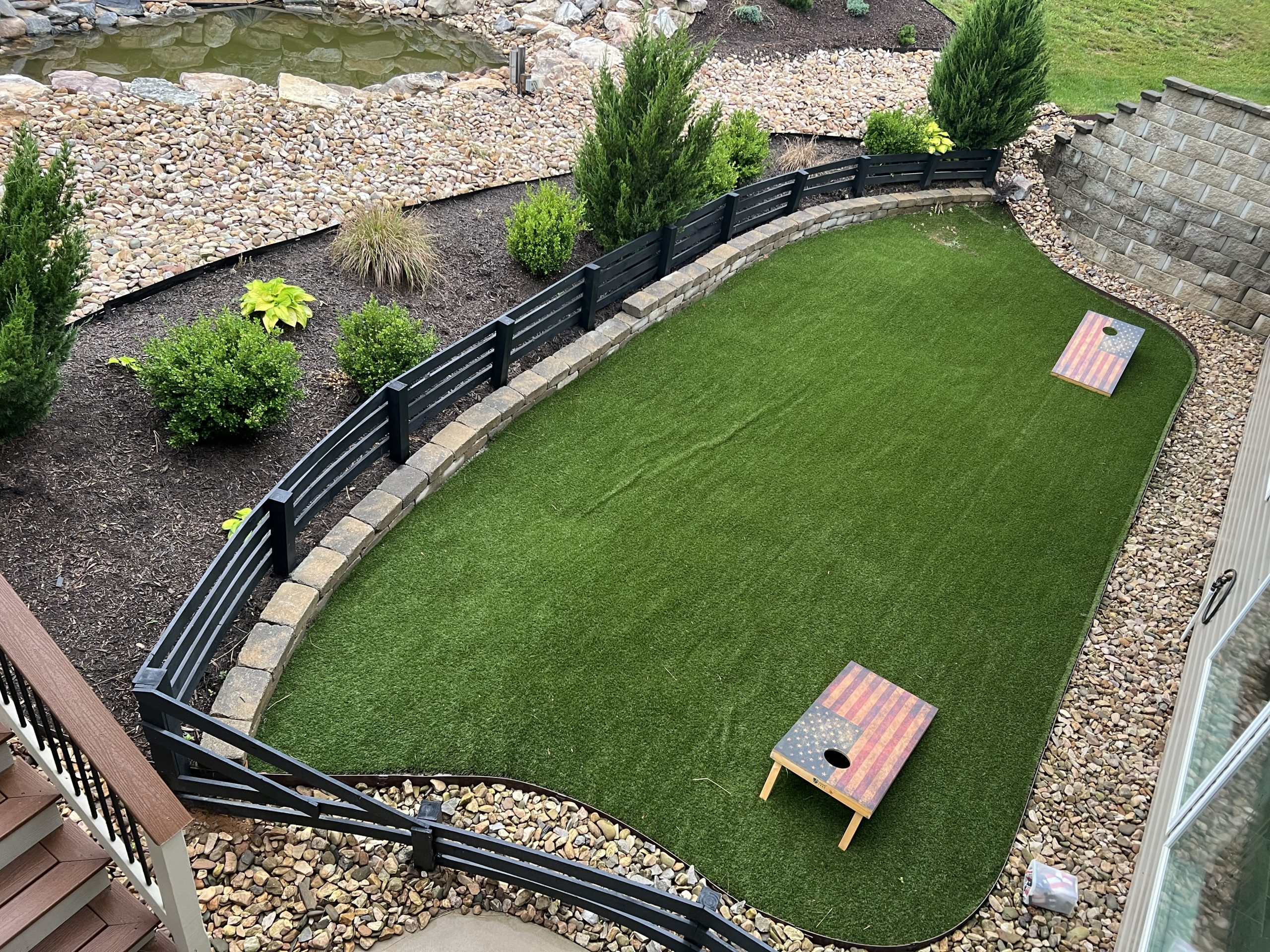 Aerial view of a small backyard with artificial turf, two cornhole boards, and surrounding garden beds with bushes and stone borders.