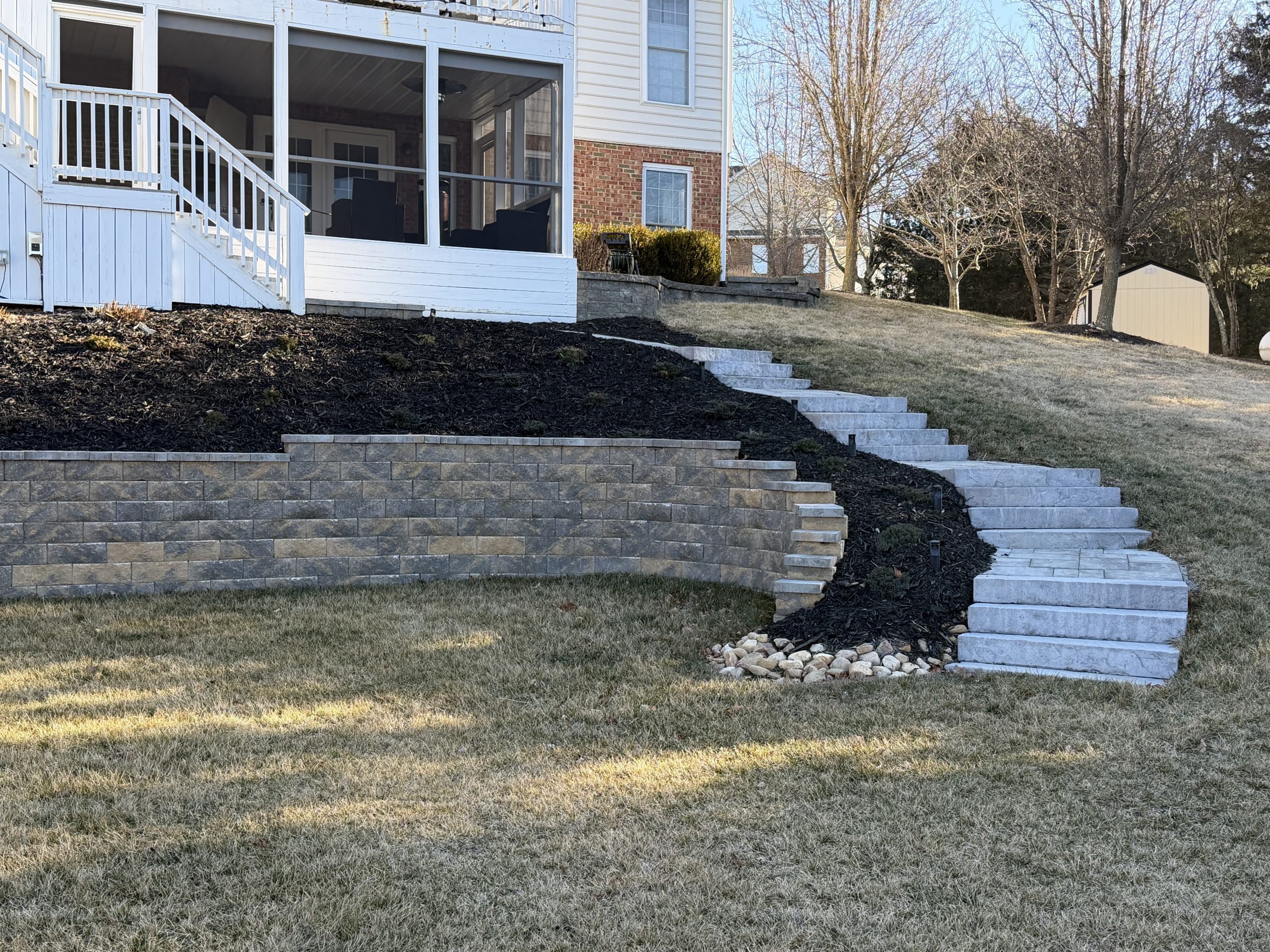 A white house with a screened porch sits atop a sloped lawn with tiered stone retaining walls and a curved stone staircase. Trees and shed in the background.